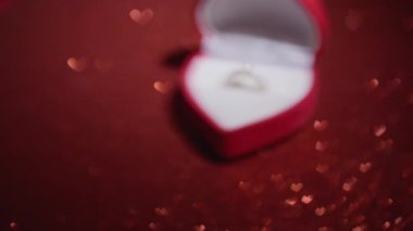 Kalp şeklinde romantik bir yüzük kutusu, içinde bir anlık bir bakış için hafifçe açık, pırıl pırıl kırmızı bir arka plana uzanır, parıldayan kalplerle, sevgililer gününde aşkı ve bağlılığı sembolize eder.