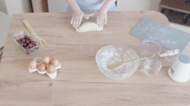 Ev mutfağında, bir kadın ahşap bir tezgahta hamur yoğururken görülüyor. Yumurta, un ve kiraz ev yapımı pişirme için hazır. Bu aşçılık hazırlığı hamur işi yapmayı da içeriyor olabilir.