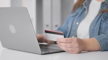 Görüntü, birbirine bağlı modern dünyamızdaki online alışveriş veya dijital ödeme sürecini net bir şekilde tasvir eden, dizüstü bilgisayar kullanırken kredi kartını sıkıca tutan bir elin yakın çekimini gösteriyor.