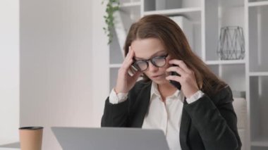 Odaklanmış bir iş kadını, dizüstü bilgisayarı üzerinde çalışırken zorlu bir telefon görüşmesi yaparken stresli görünüyor. Yoğun baskı ve çoklu görev güçlüklerinin sinyalini vermek için şakaklarını ovuyor.