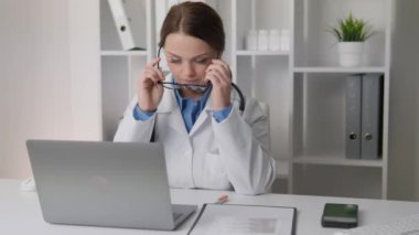 Modern bir ofiste, beyaz önlüklü ve gözlüklü stresli bir tıp uzmanı yoğun bir şekilde laptoplarına yoğunlaşarak, tam bir uzmanlık gerektiren görevlere odaklanırlar.