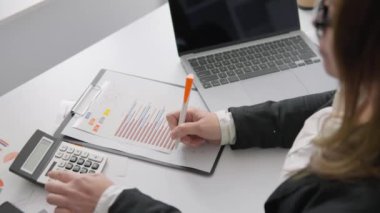 Yetenekli bir iş adamı finansal belgeleri gözden geçirir, turuncu kalemle notlar alır, hesap makinesinde rakamları hesaplar ve bilgili kararlar almak için dizüstü bilgisayardaki verileri analiz eder.