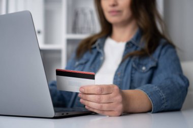 Bir kadın masasında oturuyor, dizüstü bilgisayarına odaklanıyor ve bir kredi kartı tutuyor, çevrimiçi alışveriş sitelerini keşfediyor ve alışverişler yapıyor, modern perakende teknolojisinin kolaylığını vurguluyor.