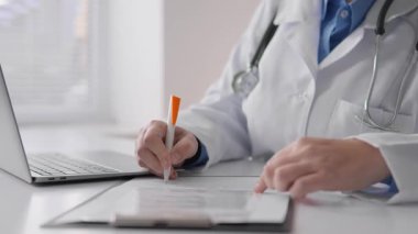 Temiz beyaz önlüklü kendini adamış bir tıp uzmanı, bir not panosuna notlar yazar ve bir dizüstü bilgisayara yazarak, klinik bir ortamda önemli bir sağlık belgelendirme sürecini gösterir.