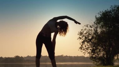 Gün batımında zarif bir şekilde yoga yapan, esnemek için ustalıkla eğilen, doğanın sunduğu dingin güzellik arasında sakinlik ve denge hissini somutlaştıran büyüleyici bir siluet.