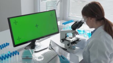 Modern bir laboratuvarda, bir bilim adamı yakın bir bilgisayardaki canlı yeşil ekranla yüksek güçlü bir mikroskop altında numuneleri inceler ve araştırmadaki hassasiyet ve gözlemin önemini vurgular.