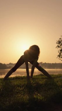 Gün batımında yoga yapan bir kadının güzel tanımlanmış silueti, vücudunu ve zihnini suyun kenarındaki sakin ve resimsel doğal manzaranın tam ortasında mükemmel bir şekilde harmanlıyor.