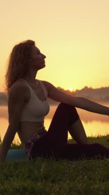 Güneş batarken, bir kadın göl kenarında yoga pratiği yapıyor, sakin bir poz veriyor, vücudunu ve zihnini mükemmel bir denge içinde harmanlarken huzur yayıyor.