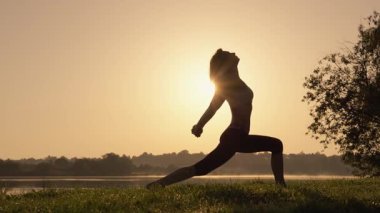 Gün batımında yoga yapan güzel ve dingin bir kadın silueti. Sıcak güneş sakin bir ufkun ardında yavaşça batarken iç huzur ve denge duygusunu somutlaştırıyor.