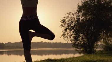 Siluetli bir figür, günbatımında sakin bir gölün yanında zarif bir yoga pozu verir, vücut ve zihin arasındaki uyumu sembolize eder, sıcaklık ışığı yansıma sırasında doğa ve iç huzuru vurgular.
