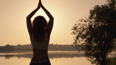 Şafak vakti yoga yapan bir bireyin silüeti. Dua eden eller, bedenle zihin arasındaki uyumu sembolize ediyor. Huzurlu bir göle karşı kurulmuş, sabahları iç huzuru yansıtıyor.