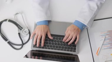 Beyaz önlüklü deneyimli bir sağlık uzmanı yakınlarda steteskopu olan bir dizüstü bilgisayarda yazarak modern tıp ve sağlık uygulamalarına teknolojinin entegrasyonunu sembolize ediyor.