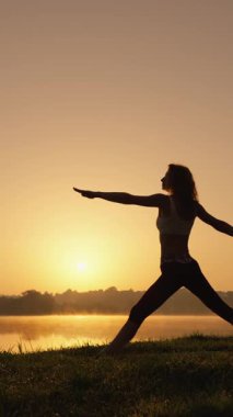 Huzurlu bir kadın silueti yoga pozisyonunda nefes kesici bir günbatımına karşı zarifçe konumlandırılmış, vücudu ve zihni mükemmel bir şekilde uyum içindeyken, sakin bir kucaklama ve güzelliğe dalmış.
