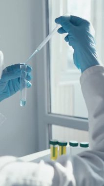 Laboratuvar önlüğü ve eldiveni giymiş kendini adamış bir bilim adamı sıvıyı test tüpüne aktarmak için pipetini kullanır. Bu da bilimsel araştırmaların ve temel aşı geliştirmenin titiz doğasını gözler önüne serer.