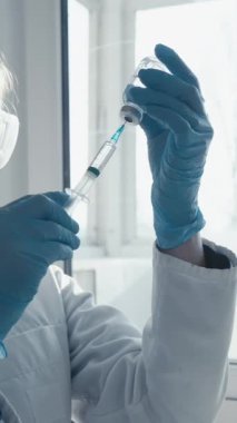 Beyaz önlüklü ve mavi eldivenli odaklanmış bir bilim adamı güneş ışığı altındaki bir laboratuvardaki bir şişeden sıvı çıkararak gerekli tıbbi araştırmaların önemini vurgulayarak bir aşı hazırlıyor.