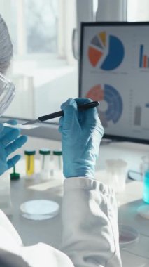 Laboratuvar önlüğü ve eldivenli bir bilim adamı hayati bir aşı araştırması yürütüyor, bir test tüpünü dikkatlice inceliyor ve bir monitörde karmaşık verileri analiz ediyor, sağlık hizmetleri için yaptıkları çalışmaların önemini vurguluyor.