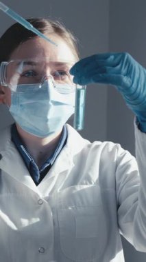 Koruyucu giysi giyen bir bilim adamı modern bir laboratuvarda bir test tüpünü inceleyerek, tıbbi araştırmalardaki gelişmeleri ve kamu sağlığı için sürdürülen önemli aşıların geliştirilmesini vurguluyor.