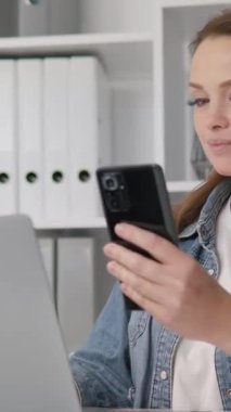 Modern bir masadaki akıllı telefonuna odaklanmış genç bir kadın, temiz, düzenli bir çalışma alanında, çekici yeşillik ve canlı bitkilerle birlikte, davetkar bir çevre sağlıyor.