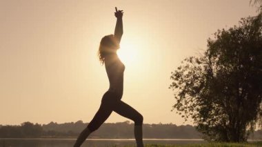 Mükemmel yoga pozu vermiş sakin ve zarif bir kadın silueti huzur dolu bir gölün üzerinde nefes kesici bir gün doğumunun çarpıcı zeminine karşı, güzel bir şekilde somutlaşmış sükunet ve ahenk içinde.