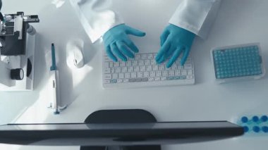Mavi eldivenli bir araştırmacının klavyede daktilo kullandığı bir laboratuvar çalışma istasyonunun tepeden tırnağa görüntüsü. Yakınlarda, odaklanmış bir bilimsel ortamı yansıtan bir mikroskop ve laboratuvar ekipmanı bulunuyor..