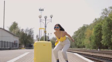 Canlı sarı bluzlu genç bir kadın ve uygun bir bavul neşeli bir tren istasyonunda taksiyi selamlıyor, tasasız bir yaz ruhunu ve yaklaşan yolculuğu için heyecanını somutlaştırıyor.