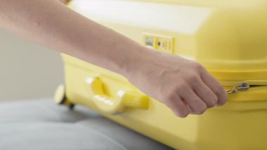 Yakın plan çekimde, parlak sarı bir valizin fermuarını dikkatlice çekerek önlerindeki heyecanlı tatil macerasına hazırlanmanın verdiği heyecanı ve beklentiyi mükemmel bir şekilde gözler önüne seriyor.