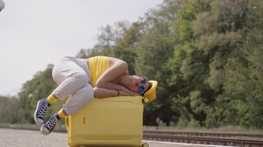 Şık ve şık yaz kıyafetleri içinde genç bir kadın parlak sarı bavulunda dinleniyor. Yeşilliklerin arasına kurulmuş heyecanlı yolculuğu başlamadan önce bu huzurlu anın tadını çıkarıyor.