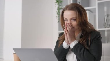 Sıkıntılı, uzun saçlı bir iş kadını laptopunun başına oturur, yoğun iş sorumluluklarından kaynaklanan yoğun endişe yüzünden yüzünü elleriyle kapatır, artan stresin ortasında odaklanmaya çalışır.