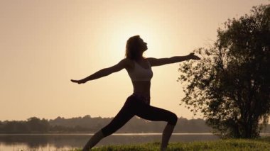 Gün batımında huzur dolu bir gölün kenarında yoga yapan güzel bir kadın silueti, alacakaranlıkta büyüleyici bir şekilde parlayan ufka karşı mükemmel bir zarafet ve sükunet simgeliyor.