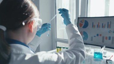 Laboratuvar önlüğü ve gözlüklü kendini adamış bir bilimadamı renkli test tüpleri ve verileri olan parlak ışıklı bir laboratuvarda bir pipetle yenilik ve keşif atmosferi yaratıyor.