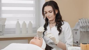 Yetenekli bir kozmetik uzmanı, bir kozmetik prosedürü sırasında müşterinin yüzüne serumu uyguluyor. Rahatlamayı ve sağIığı destekleyen sakin bir klinikte profesyonel cilt bakımını gösteriyor.