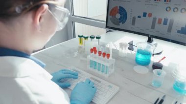 Bir laboratuvar ortamında kendini adamış bir bilim adamı titizlikle test tüpleri ile çalışır ve bir bilgisayar ekranında veri analizini izler, tıptaki ileri bilimsel araştırmaları yansıtır..