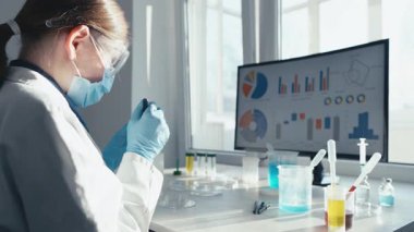 Laboratuvar önlüğü ve koruyucu giysileri içinde kendini adamış bir kadın bilim adamı, renkli sıvılar ve veri görselleştirmeleriyle çevrili iyi donanımlı bir tıp laboratuarında örnekleri titizlikle analiz ediyor..
