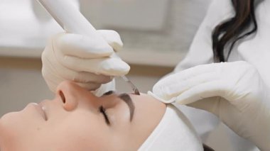 Tanınmış ve saygı duyulan bir klinikte çok yetenekli ve deneyimli bir kozmetik uzmanı, geniş kapsamlı ve detaylı bir cilt tedavisi sırasında müşterinin alnına bir cihaz takıyor.