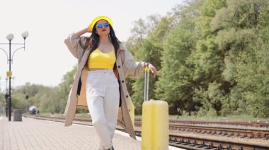 Parlak sarı bir şapka ve son moda güneş gözlüğü takan şık genç bir kadın kırsal tren raylarında geziniyor, yaz yolculuğuna canlı bir bavulla hazır, bu sezonun macerasını somutlaştırıyor.