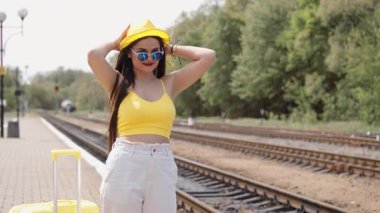 Şık, genç bir kadın, parlak sarı bir kıyafet ve şık bir şapkayla yoğun bir tren istasyonunda duruyor. Bavuluyla yaz tatiline hazırlanıyor. Yeni bir maceranın heyecanını kucaklıyor.