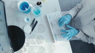 Laboratuvar önlüğü ve eldiven giyen kendini işine adamış bir bilim adamı, araştırma ve deneyler için bir monitörde karmaşık verileri analiz eden petri kabı ve çözümler gibi laboratuvar ekipmanlarıyla çevrili bir bilgisayarda çalışıyor.