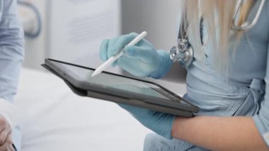 Eldivenli bir sağlık uzmanı, bir konsültasyon sırasında hasta bilgilerini gözden geçirmek için bir tablet kullanıyor ve günümüz sağlık hizmetlerinde modern tıp teknolojisinin gelişmelerini gösteriyor.