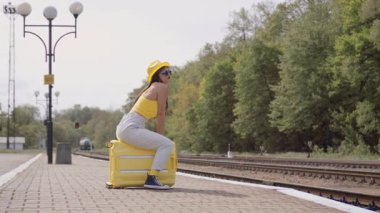 Parlak giyinmiş genç bir kadın, güneşli bir tren istasyonunda parlak sarı bir bavula oturur, yaz yolculuğunun kaygısız ruhunu somutlaştırarak sabırla yaklaşan macerasını bekler.