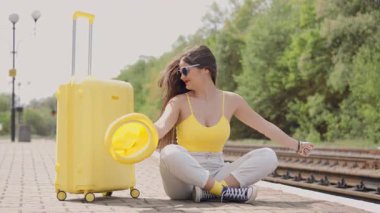 Parlak sarı bluzlu ve son moda güneş gözlüklü hayat dolu genç bir kadın yoğun bir tren istasyonunda neşeli sarı bavulunun yanında oturur ve yaz tatilinin ve maceranın neşesini yakalar.