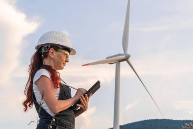Sert bir şapka takan kendini adamış bir teknisyen, yenilenebilir enerji çözümleriyle ilgili performans ölçümlerini analiz etmek ve değerlendirmek için titizlikle bir tablet kullanan bir rüzgar türbininin yanında duruyor.
