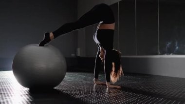 Bir kadın dengeli bir topun üzerinde etkileyici bir şekilde amuda kalkar, esnekliğini, gücünü ve sanatını minimalist fitness ortamında sergiler, atletizmin zarafetini ve zarafetini vurgular.