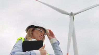 Güvenlik teçhizatı ve pano ile donatılmış bir kadın mühendis, bulutlu bir günde bir rüzgar türbini değerlendirerek, sürdürülebilir bir gelecek için hayati önem taşıyan yenilenebilir enerjideki gelişmeleri vurguluyor.