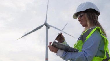 Yetenekli bir kadın rüzgar enerjisi teknisyeni tabletindeki karmaşık verileri, yenilenebilir enerji ve alandaki gelişmeleri vurgulayan yüksek türbinlerin yanında analiz ediyor.