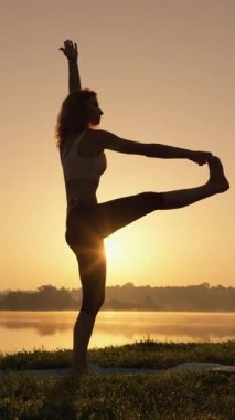 Şafak vakti yoga yapan güzel, dingin bir kadın silueti, doğa tarafından çevrelenmiş, sakin sularda huzur ve sükunetin vücut bulmuş hali.