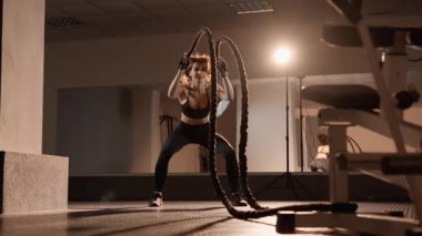 Motive olmuş bir kadın, prestijli bir spor salonunda yoğun bir savaş halatı egzersizi yapıyor. Gücünü ve konsantrasyonunu gösterirken, dayanıklılığını ve dayanıklılığını arttırıyor.