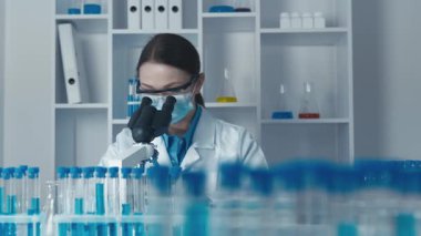 Kendini adamış bir kadın bilim adamı, renkli test tüpleri ve temel laboratuvar ekipmanlarıyla çevrili mikroskopla örnekleri analiz ederek, alanındaki araştırma ve yeniliklerin önemini vurguluyor.