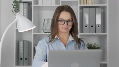 Yeşillik ve organize dosyalarla dolu canlı bir ofiste, gözlüklü genç bir kadın dizüstü bilgisayarı üzerinde çalışırken gülümsüyor, modern, üretken ve motive edici çalışma ortamını yansıtıyor.