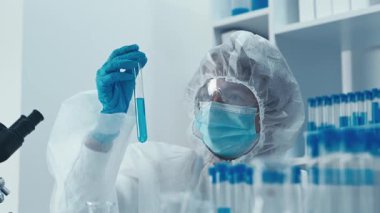 Koruyucu giysi giyen bir bilim adamı modern bir laboratuvarda parlak mavi sıvıyla bir test tüpünü inceleyerek araştırma, keşif ve teknolojide ilerlemenin hayati önemini vurguluyor.