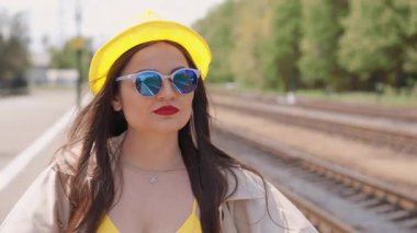 Uzun saçlı, parlak sarı elbiseli ve şık güneş gözlüklü genç bir kadın, yoğun bir tren istasyonunda duruyor, heyecan verici yaz tatiline çıkmaya hazır, olasılıklarla dolu.